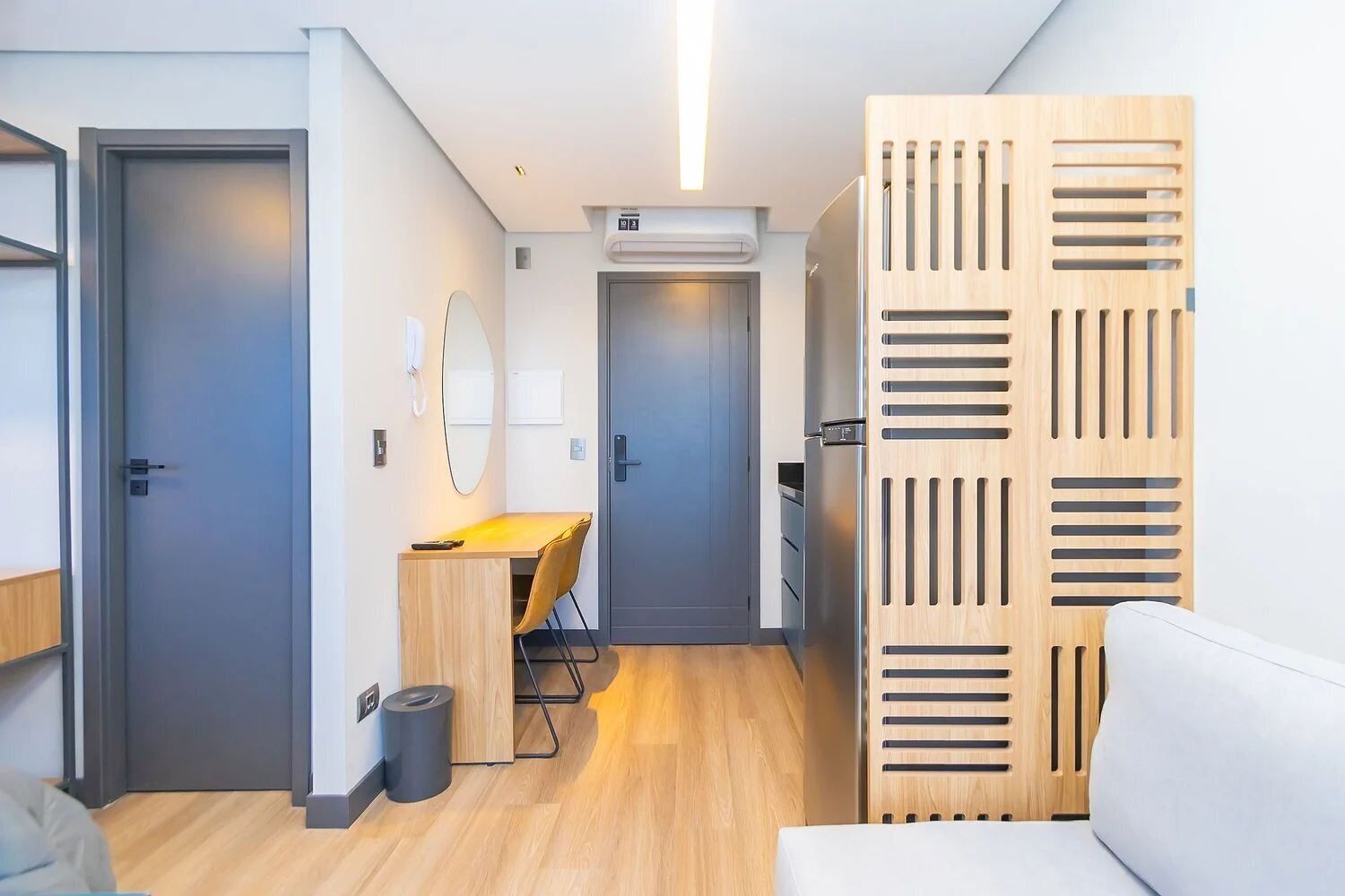 A modern hotel room with a wooden desk, a chair, a small trash bin, and a wardrobe with a unique design.