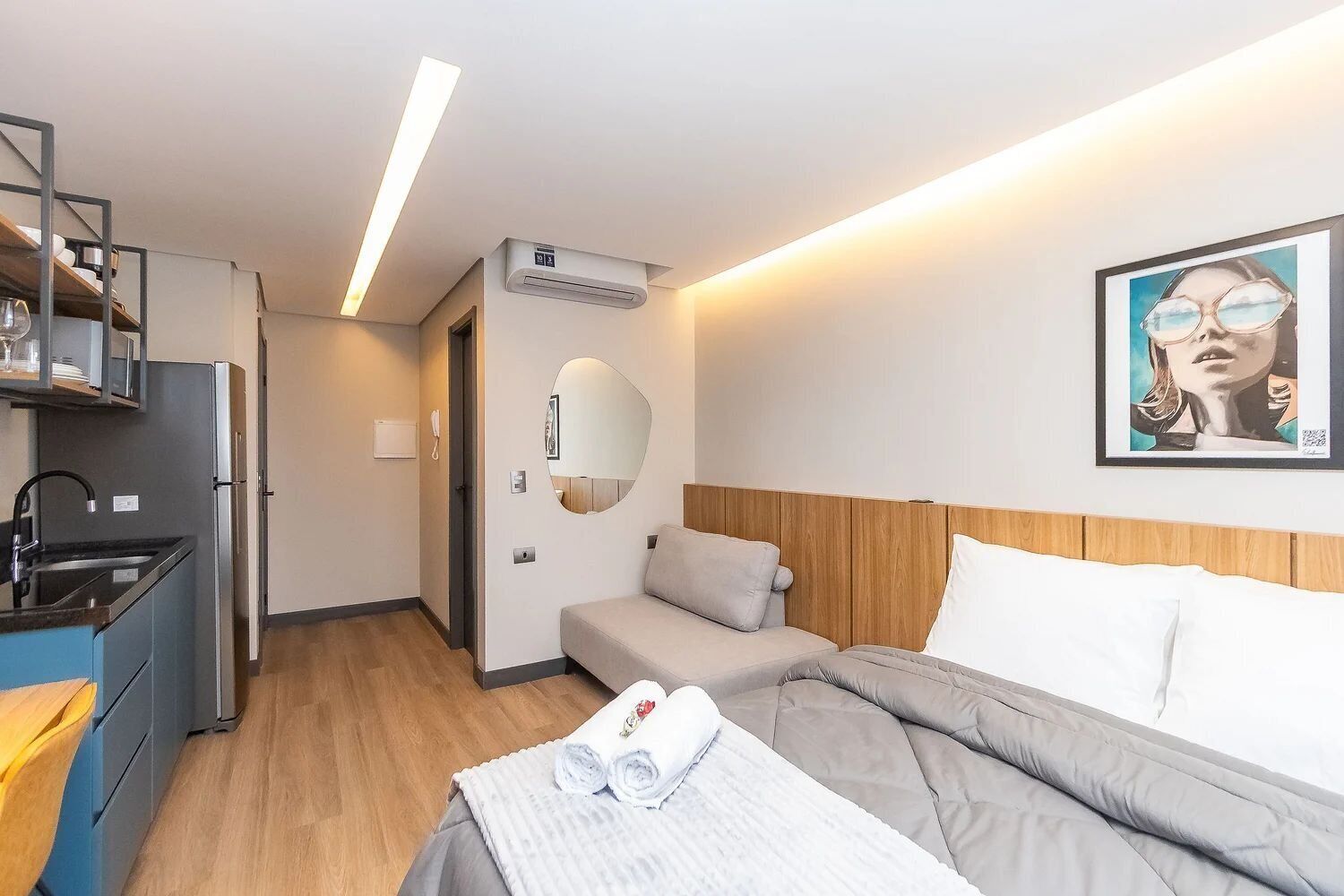 Habitación de hotel con kitchenette, una cama con cabecera de madera, un espejo redondo y un cuadro en la pared.