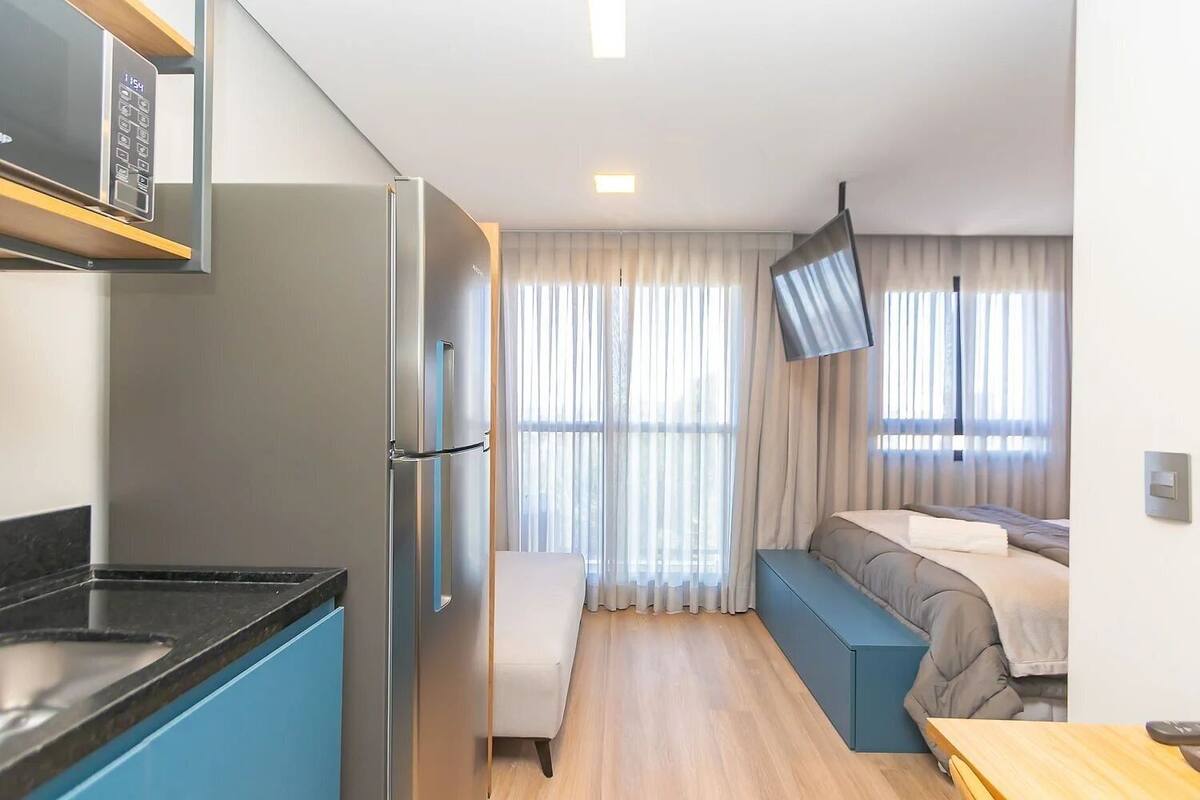 Une chambre d’hôtel compacte, dotée d’une kitchenette, d’un lit avec une banquette et d’une grande fenêtre avec des rideaux.