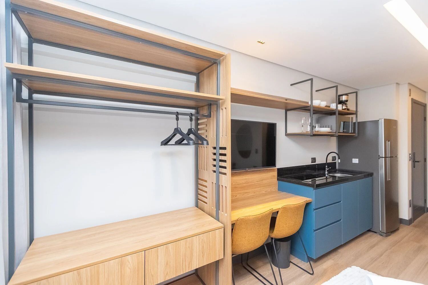 A compact kitchen with a built-in stove, a sink, and a refrigerator. There is a wooden bench and a chair, and a TV mounted on the wall.