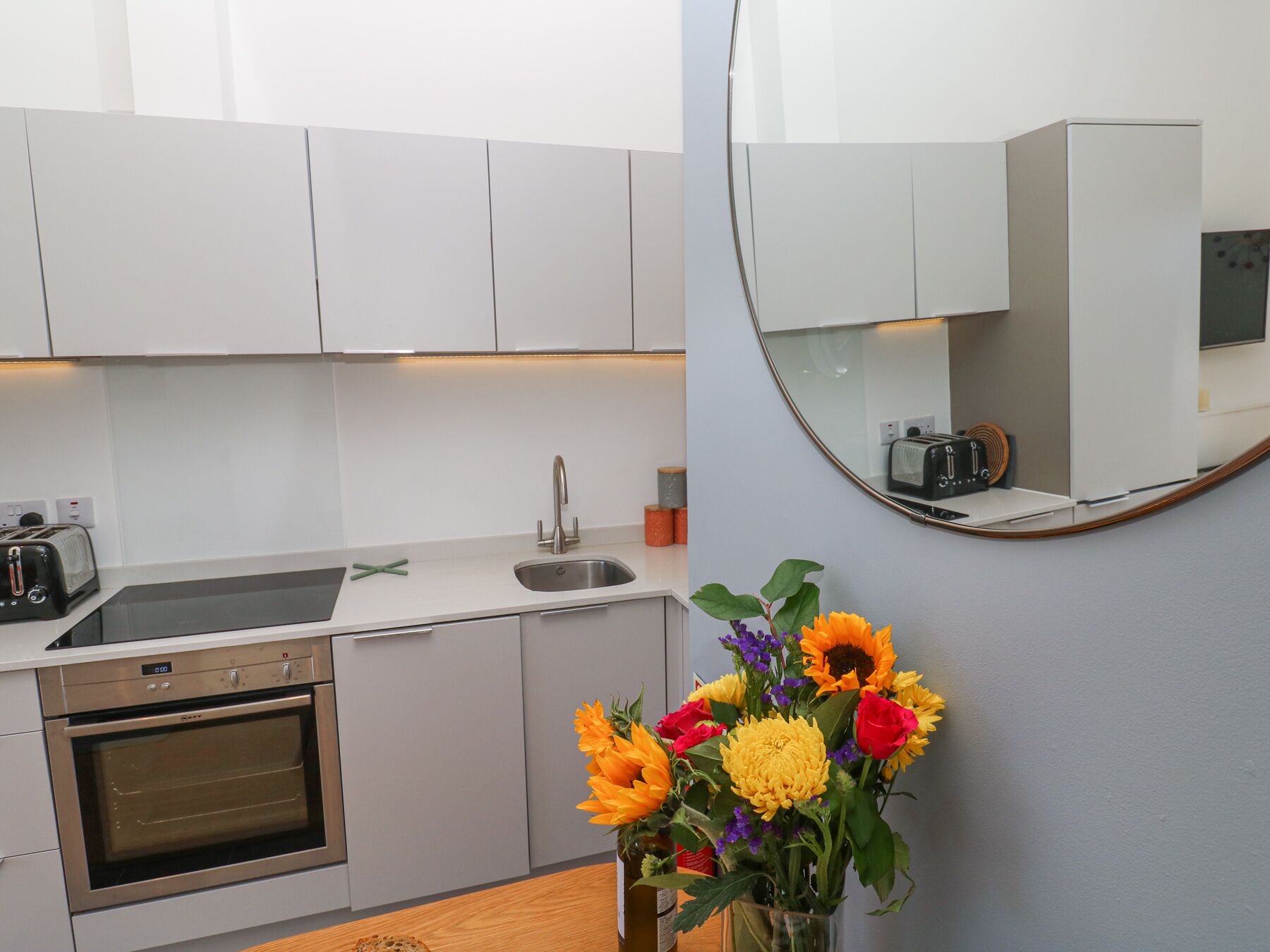 A modern kitchen with white cabinets, a stainless steel oven, a sink, and a toaster. There is a vase with flowers on the counter.
