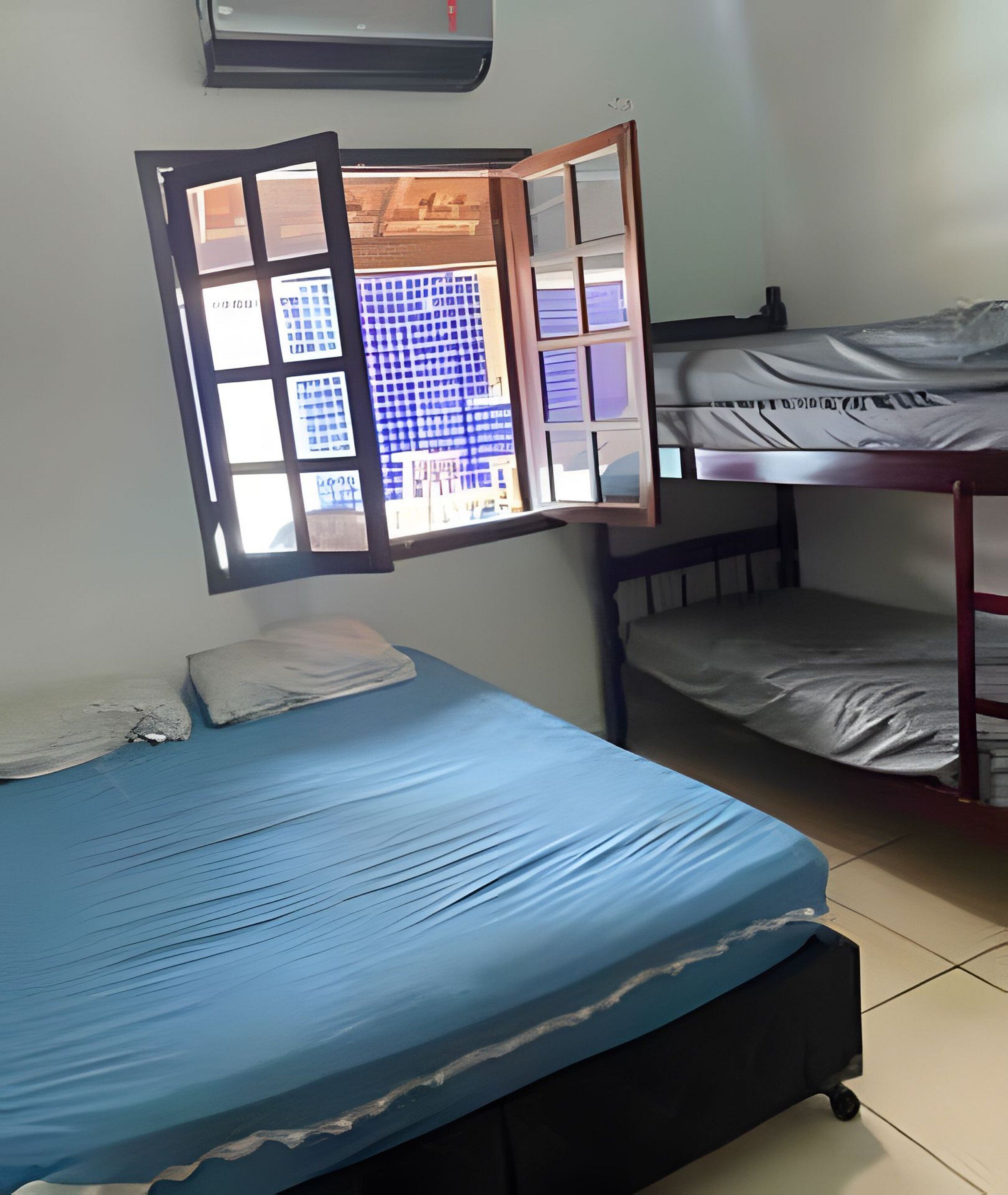 A dormitory room with a bunk bed, a single bed, and a window with a view of buildings.
