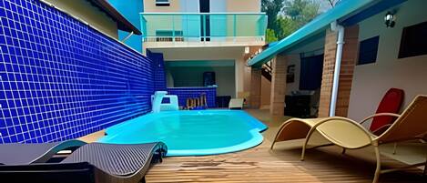 Zona de piscina con un muelle de madera, sillones de descanso y un edificio de dos pisos al fondo.