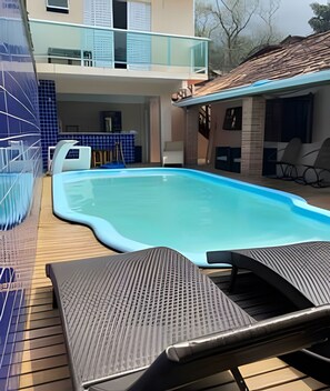 Ein Schwimmbecken mit Liegestühlen, eine überdachte Terrasse und im Hintergrund ein Gebäude mit Balkon.