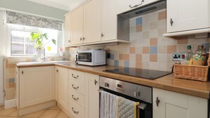 A modern kitchen with white cabinets, a microwave, an oven, and a window with a plant. - Rose Cottage (Cottingham)