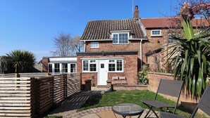 A two-story house with a brick exterior, a tiled roof, and a front door. There is a small patio area with a table and chairs, and a wooden fence surrounding the property. - Rose Cottage (Cottingham)