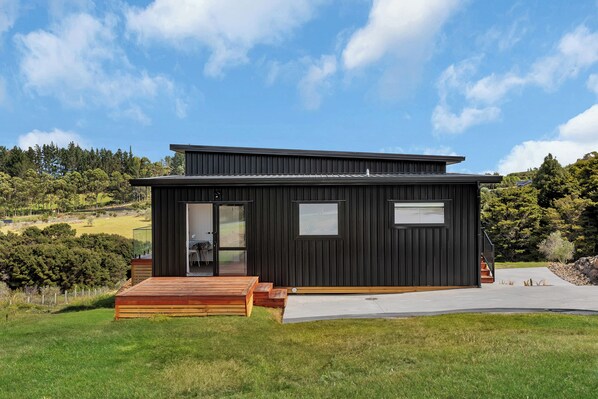 A modern black building with a wooden deck and a glass door. - Central Couples Retreat - Mangawhai Heads Unit (Mangawhai)