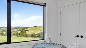 Ein Zimmer mit großem Fenster und Blick auf eine grüne Landschaft, ein kleiner Tisch mit kariertem Tischläufer und ein weißer Stuhl.