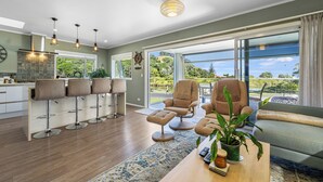 A modern living room with a large window, a sofa, a coffee table, and a bar area with stools. - The Lighthouse - Te Mata Holiday Home (Thames)