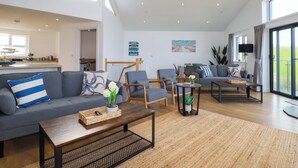 A spacious living area with a high ceiling, a kitchen in the background, and a large sliding glass door to the right. - Jacana (Perranporth)