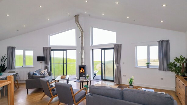 A spacious living room with a high ceiling, large windows, a fireplace, and a view of the outdoors. - Jacana (Perranporth)