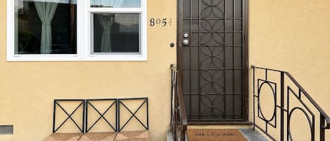 A building entrance with a number '8054' above a door, a bench outside, and a 'WELCOME' mat at the door.