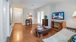 A living room with hardwood floors, a sofa, a coffee table, a TV on a wooden stand, and a door leading to another room.