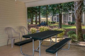 An outdoor patio with a picnic table and benches, surrounded by trees and a building in the background. - Ocean Keys 2 Blocks To The Ocean (North Myrtle Beach)