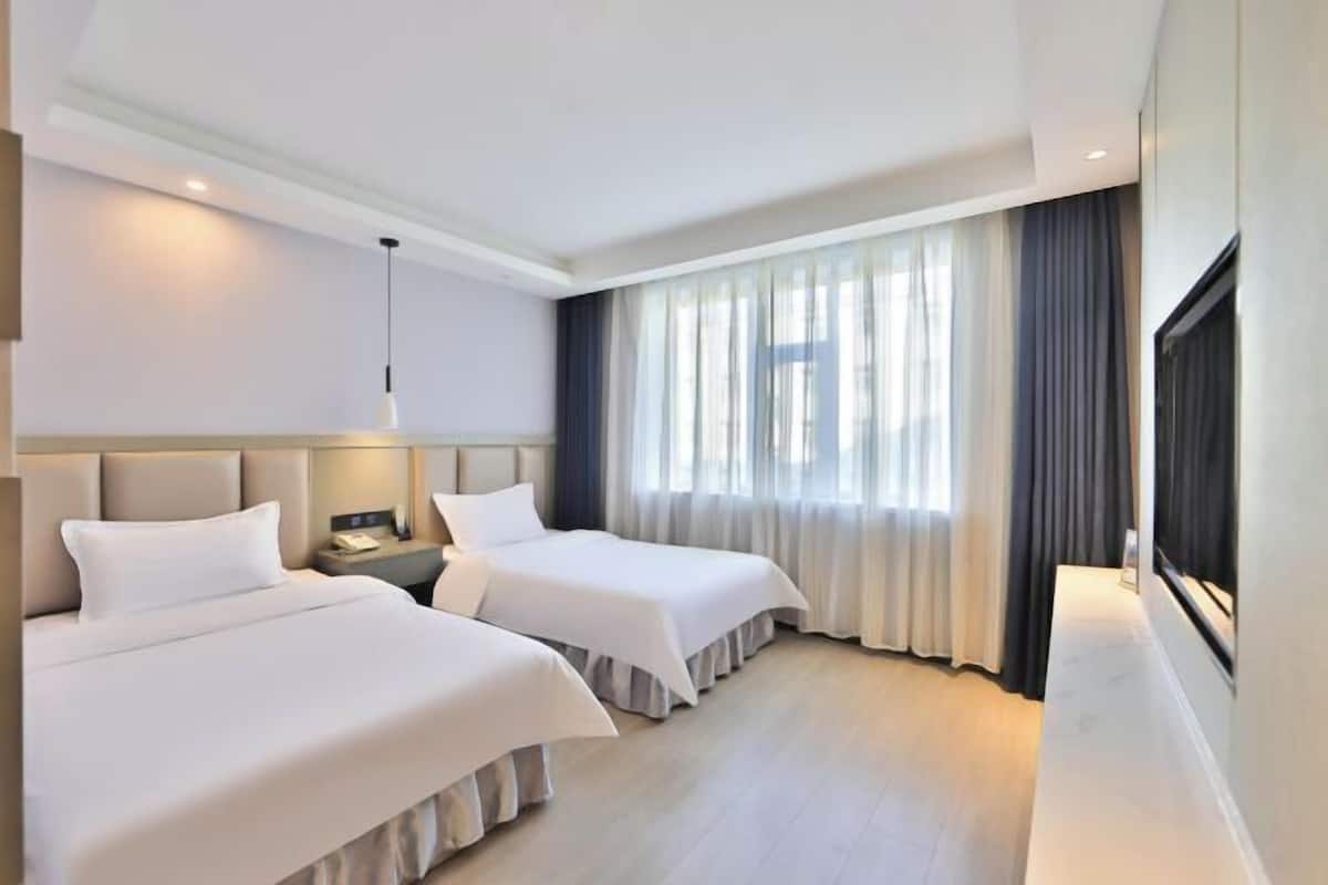 A hotel room with two beds, a nightstand, a lamp, a large window with curtains, and a view of the outside.