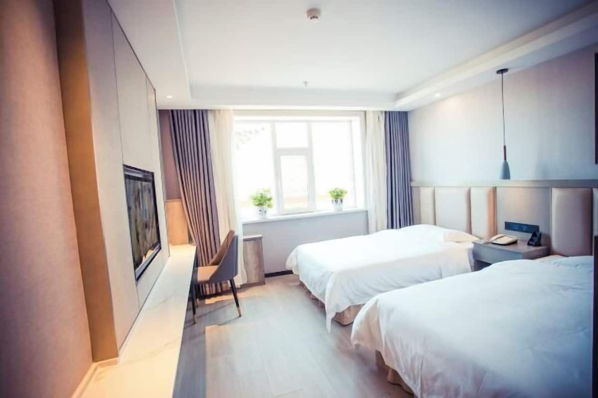 A hotel room with two beds, a desk, a chair, a window with curtains, and a bedside table with a lamp.
