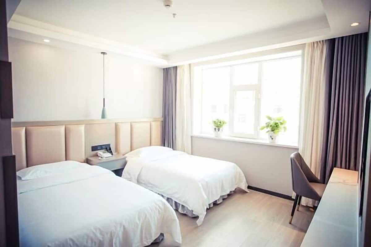 A hotel room with two beds, a desk, a chair, a window with curtains, and a plant on the windowsill.
