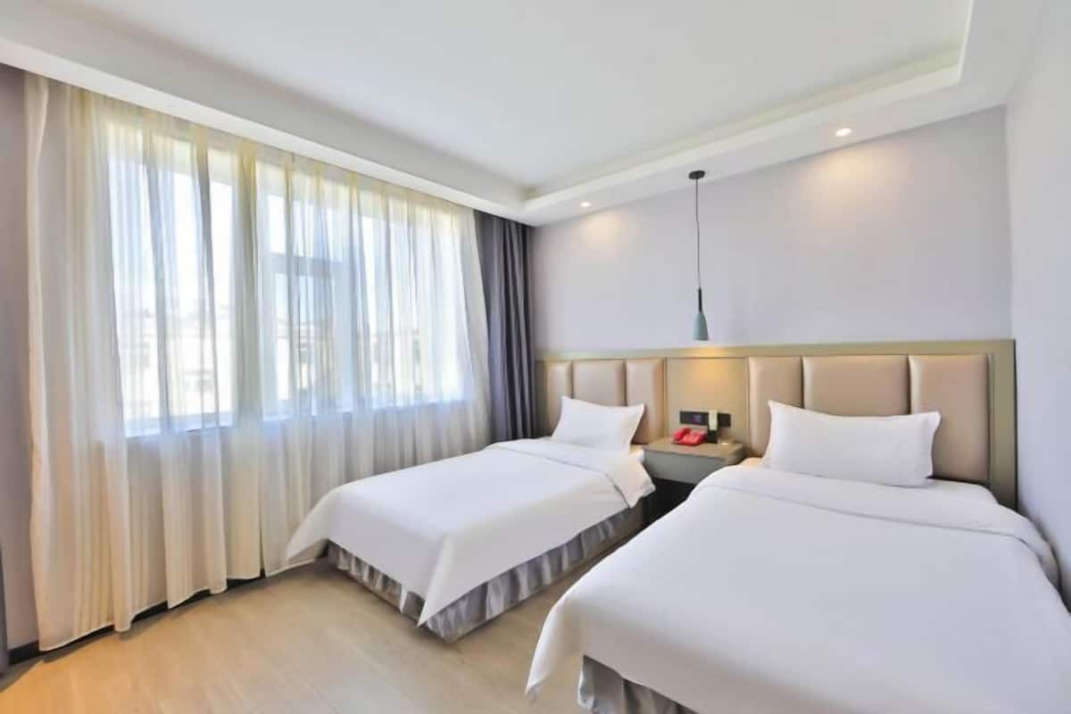 A hotel room with two beds, a large window with sheer curtains, a bedside table with a lamp, and a modern headboard.