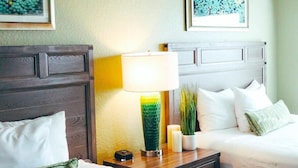 Ein Hotelzimmer mit Holz-Kopfteil, einem Nachttisch mit Lampe, einer Vase mit Pflanzen und einem Bett mit weißer Bettwäsche.