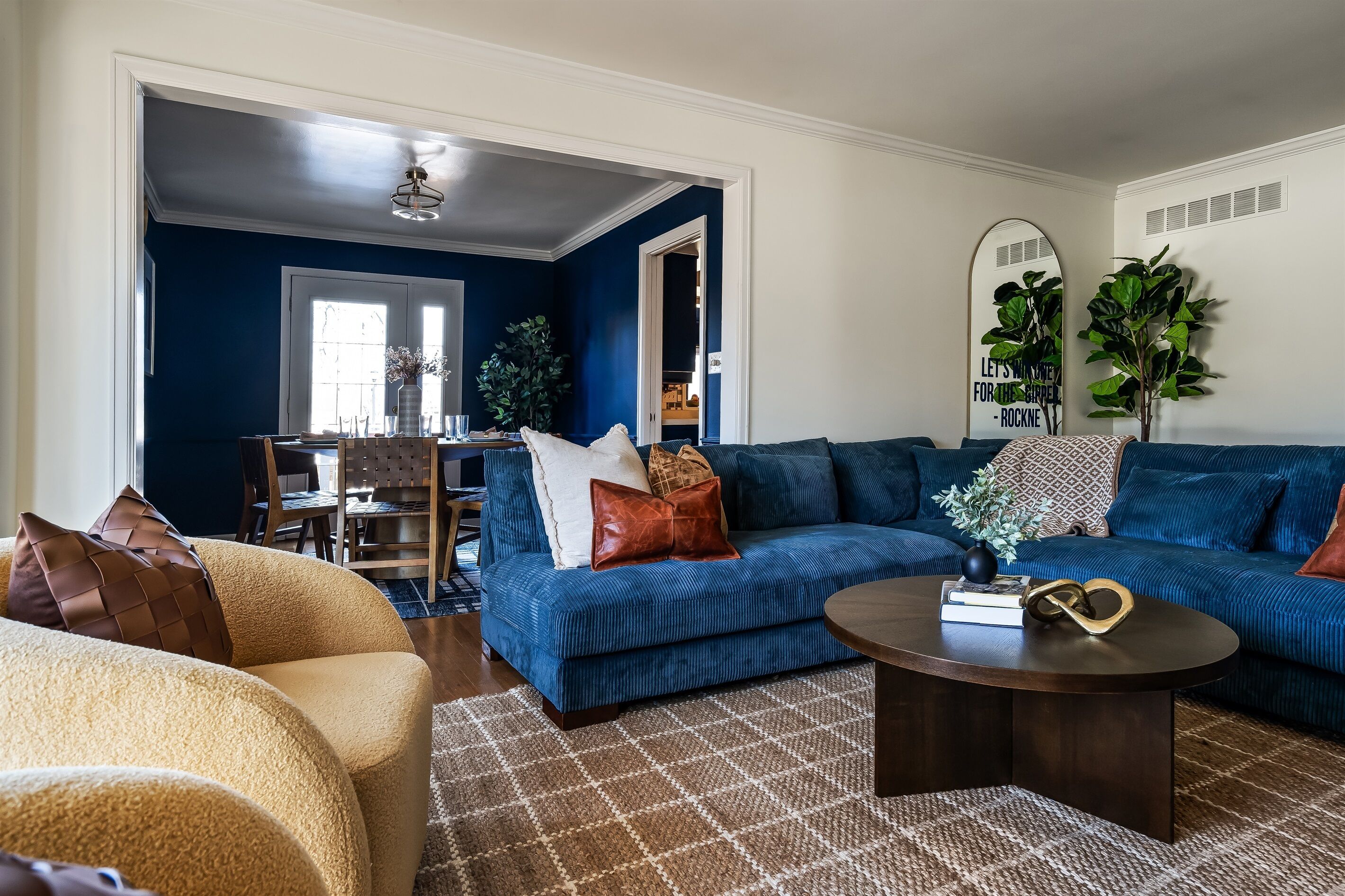 A living room with a blue sofa, a round coffee table, and a beige armchair. There is a dining area in the background with a table and chairs.