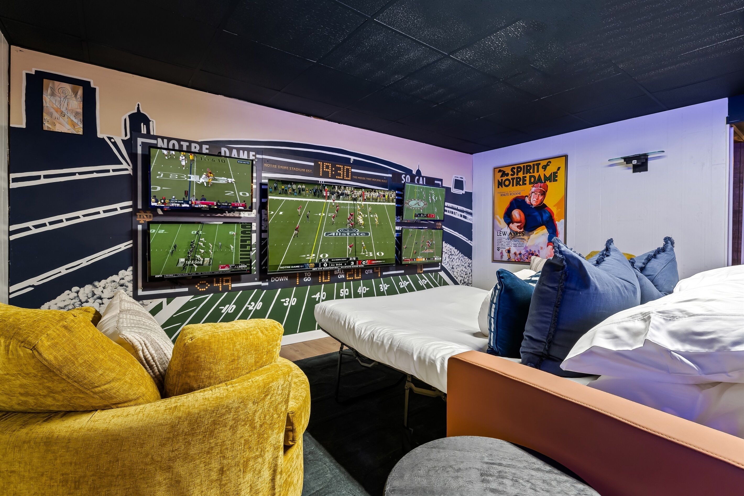 A room with a bed, a yellow armchair, and a wall-mounted TV displaying a football game.
