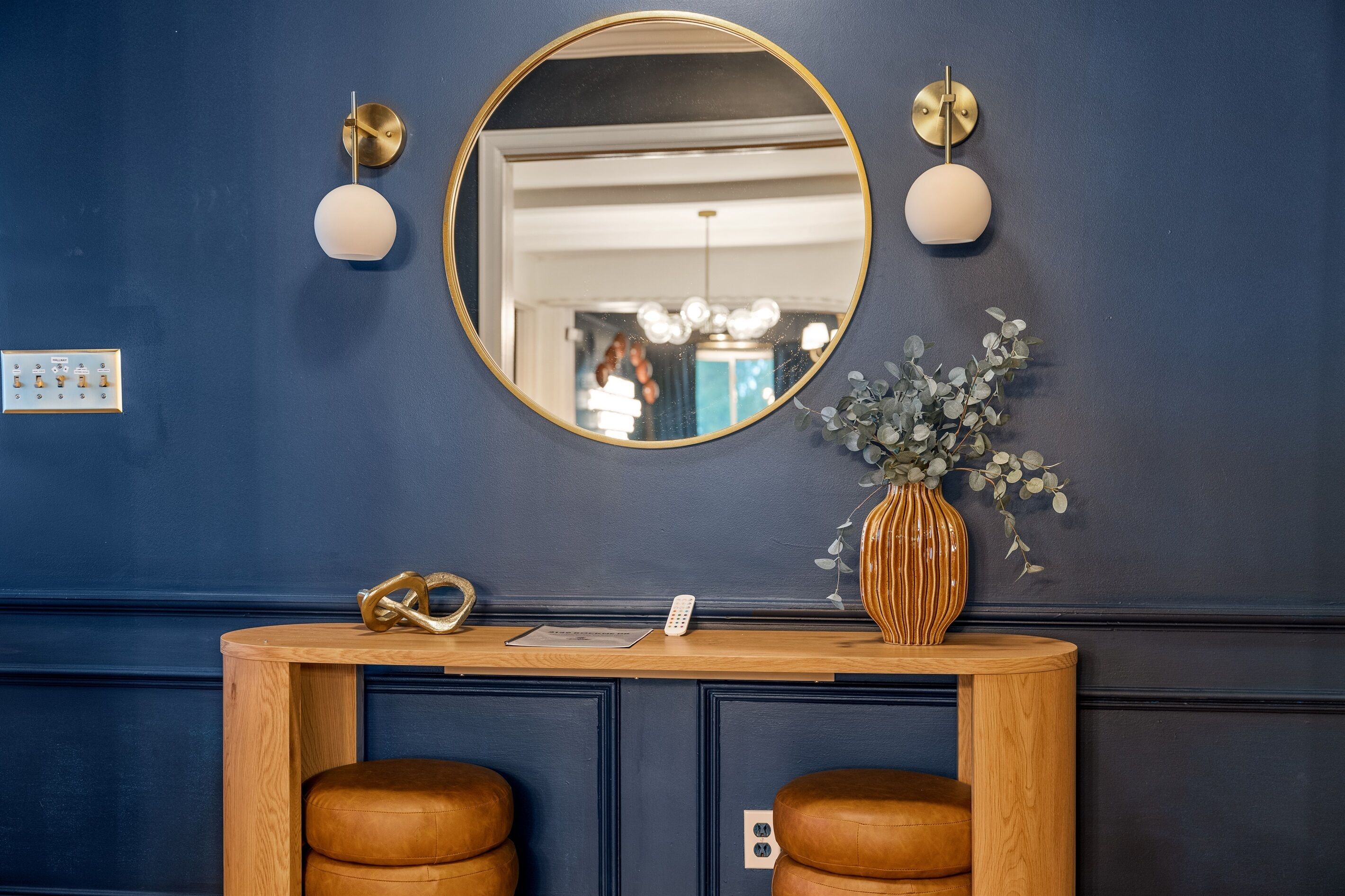 A dark blue wall with a round mirror, two wall sconces, a wooden console table with a vase and a remote control, and two leather stools.
