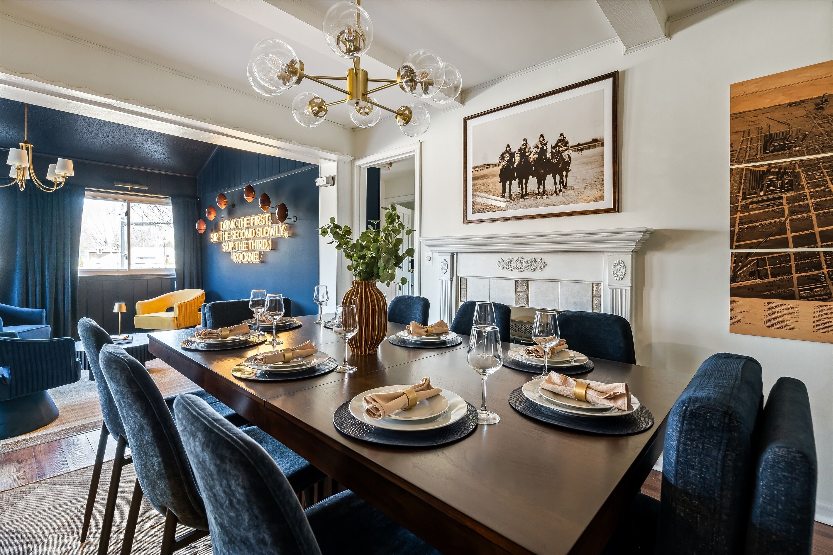 A dining area with a long table set for six, blue upholstered chairs, a chandelier, and a framed photograph on the wall.