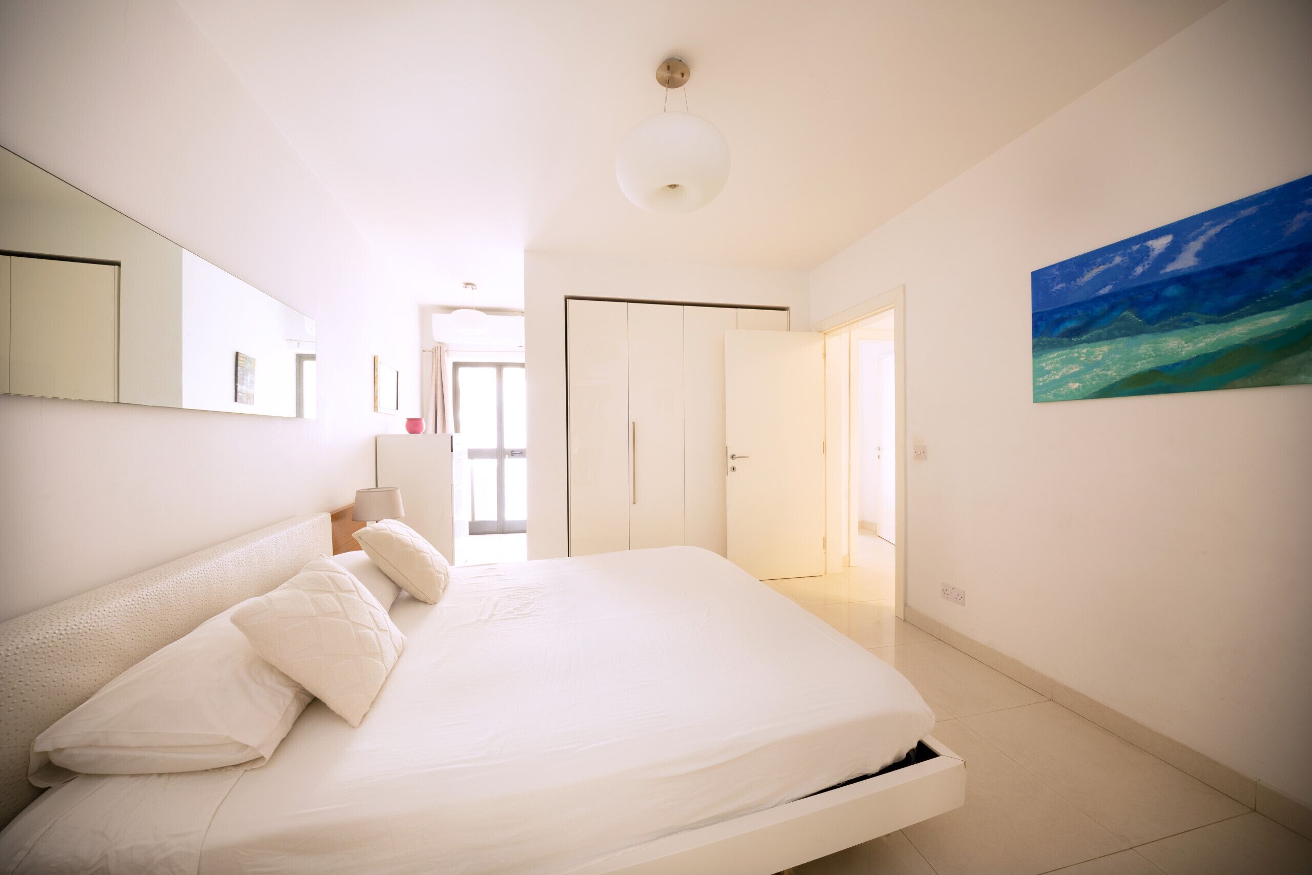A bedroom with a large bed, a bedside table, a mirror, a painting, and a wardrobe.