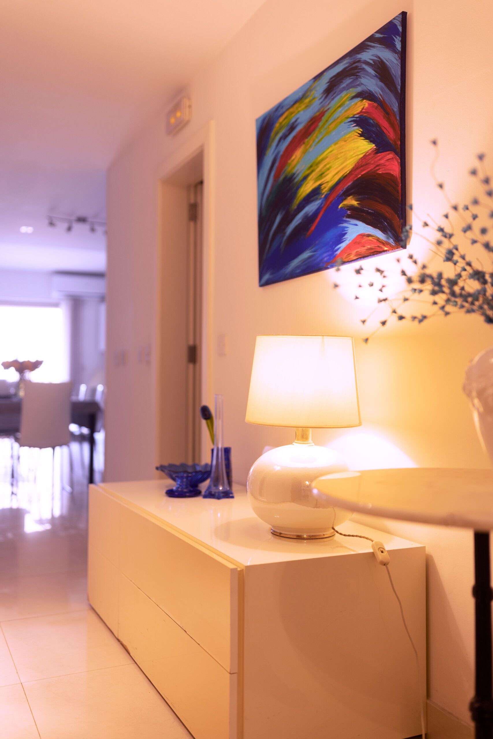 Une chambre d’hôtel moderne avec une table de chevet blanche, une lampe et un tableau abstrait aux couleurs vives accroché au mur.