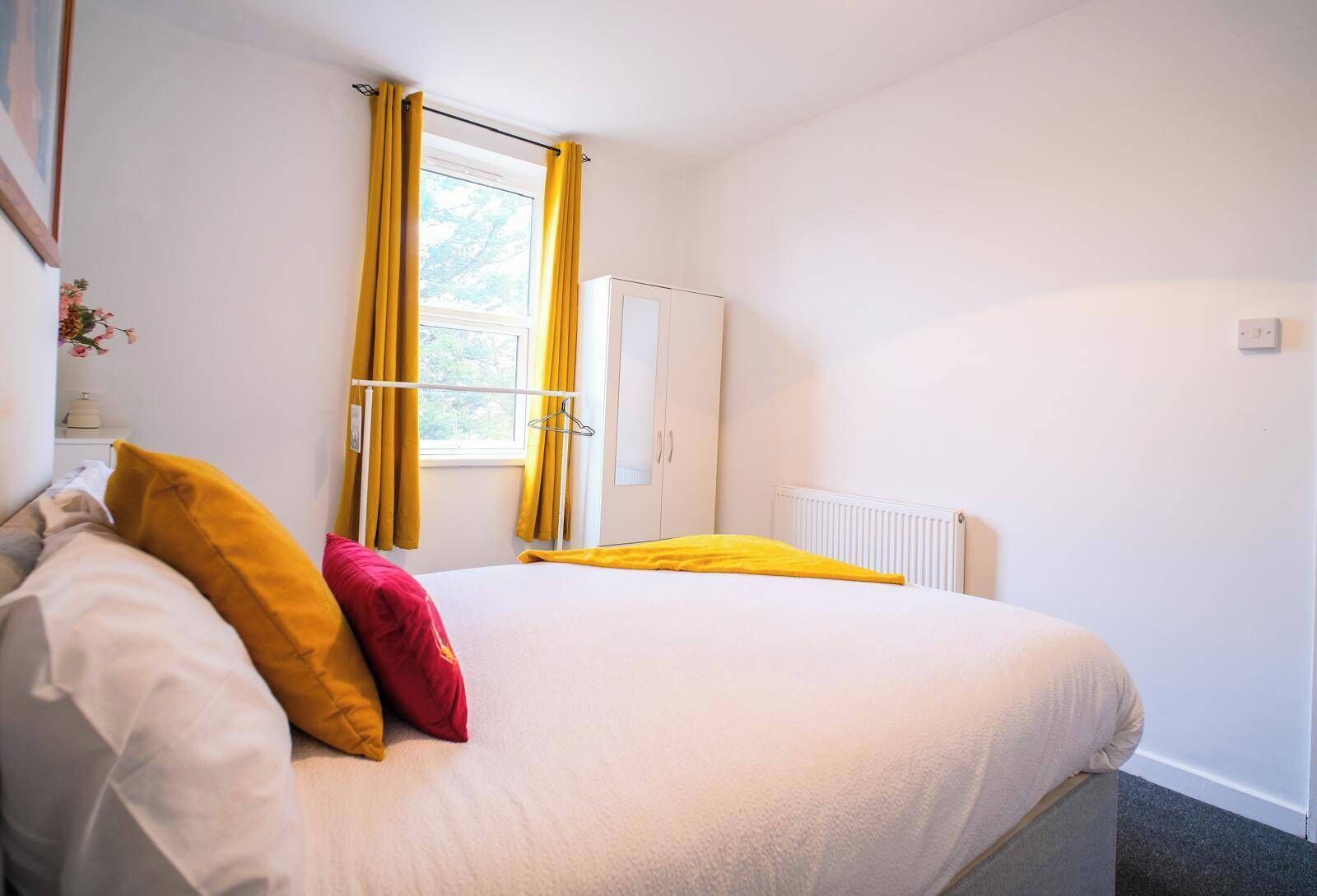 Een slaapkamer met een bed, gele gordijnen, een radiator en een witte kledingkast.