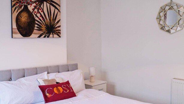 A neatly made bed with white linens, a red pillow with the word "COSY," a decorative wall piece, and a geometric wall decoration.
