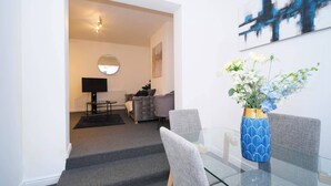 A modern living room with a glass dining table, grey sofas, a round mirror, and a blue vase with flowers.