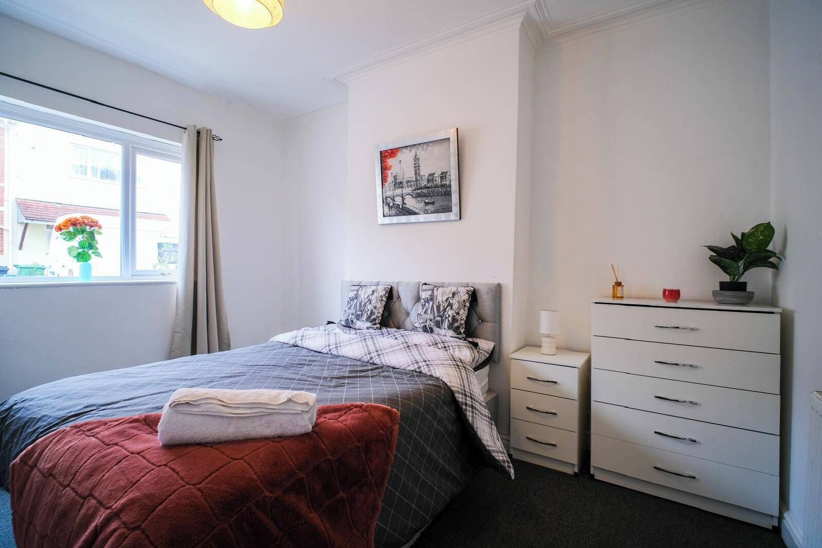 Une chambre à coucher comprenant un lit, une commode, une table de chevet, une fenêtre avec des rideaux et un tableau accroché au mur.