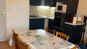 Une cuisine compacte avec des meubles de couleur sombre, un four à micro-ondes et une cuisinière. Une table à manger, recouverte d’une nappe à motifs, est entourée de chaises.