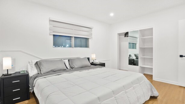 A bedroom with a bed, bedside tables, a nightstand, a window with blinds, and a built-in closet with shelves.
