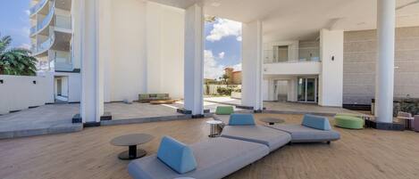 A modern outdoor lounge area with seating and tables, large pillars, and a view of a building in the background.