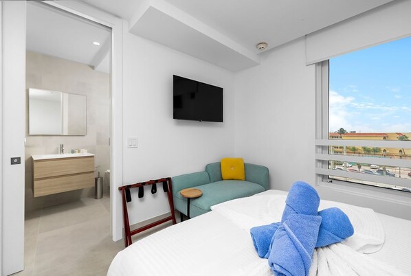Une chambre d’hôtel moderne équipée d’un lit, d’un téléviseur fixé au mur, d’une petite table, d’un canapé avec un coussin jaune et d’une fenêtre donnant sur la ville.
