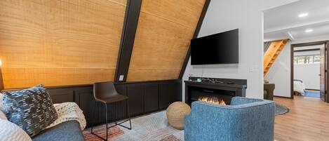 Una sala de estar moderna con chimenea, un televisor de pantalla plana montado en la pared y una zona de estar acogedora con un sillón azul y un sofá.