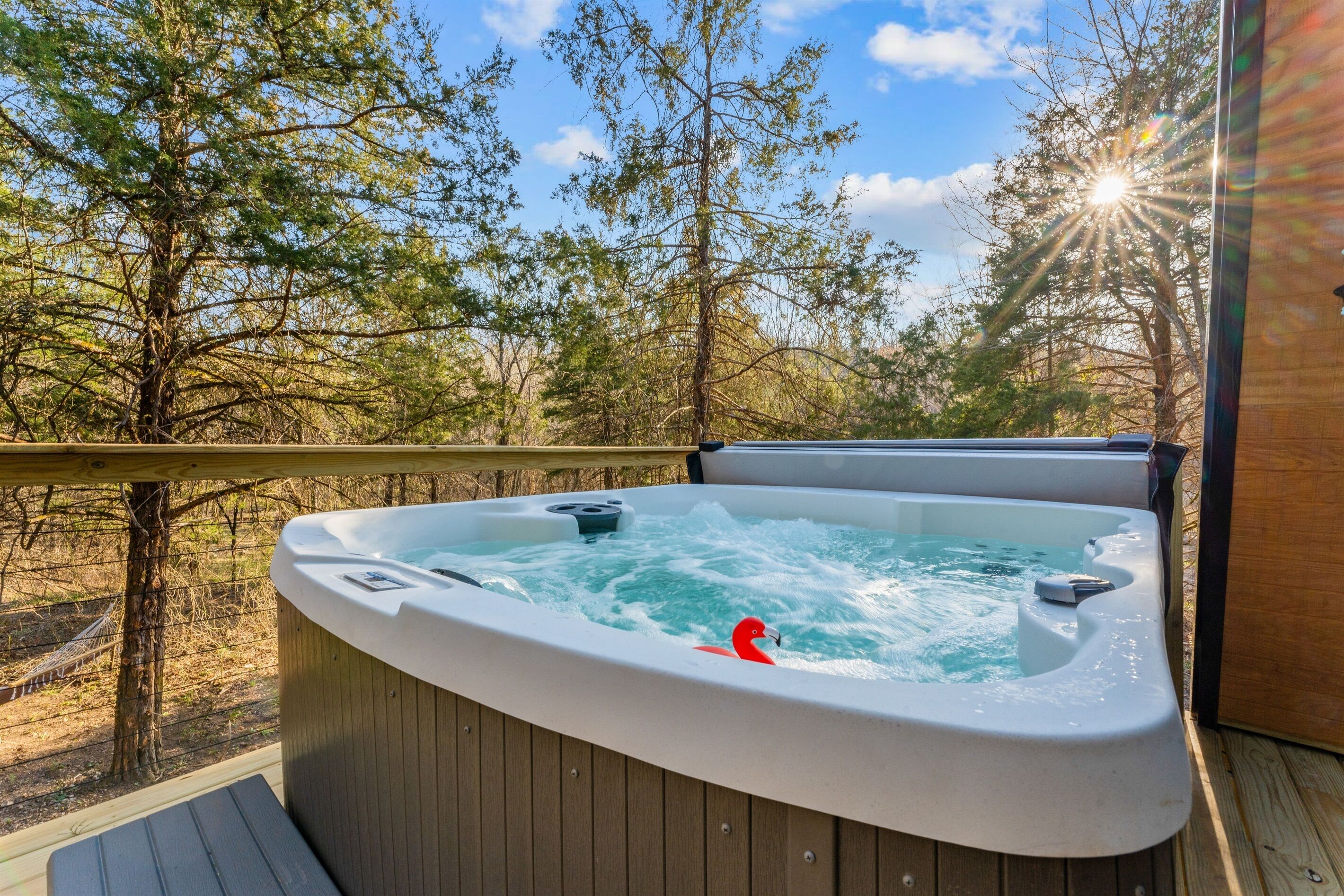 A hot tub on a wooden deck with a wooden railing, surrounded by trees and a clear sky.