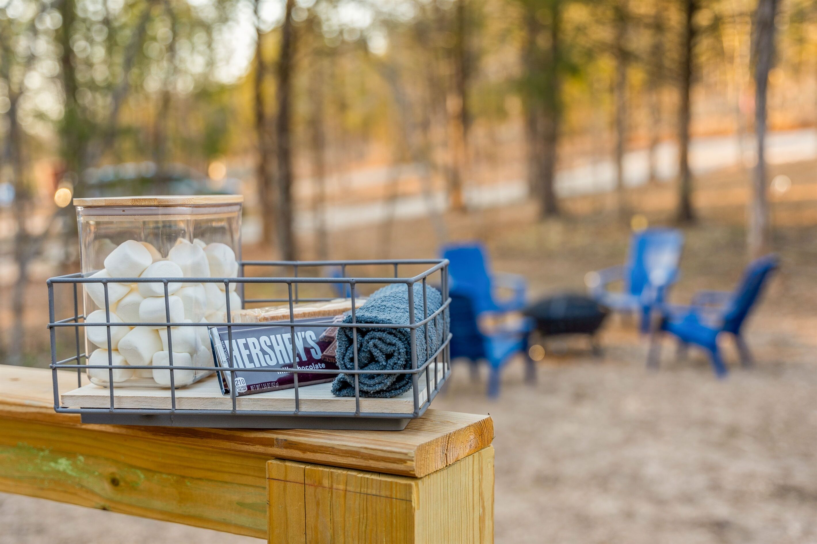 A wooden bench with a metal basket containing marshmallows and a chocolate bar, set against a forest backdrop with blue chairs.