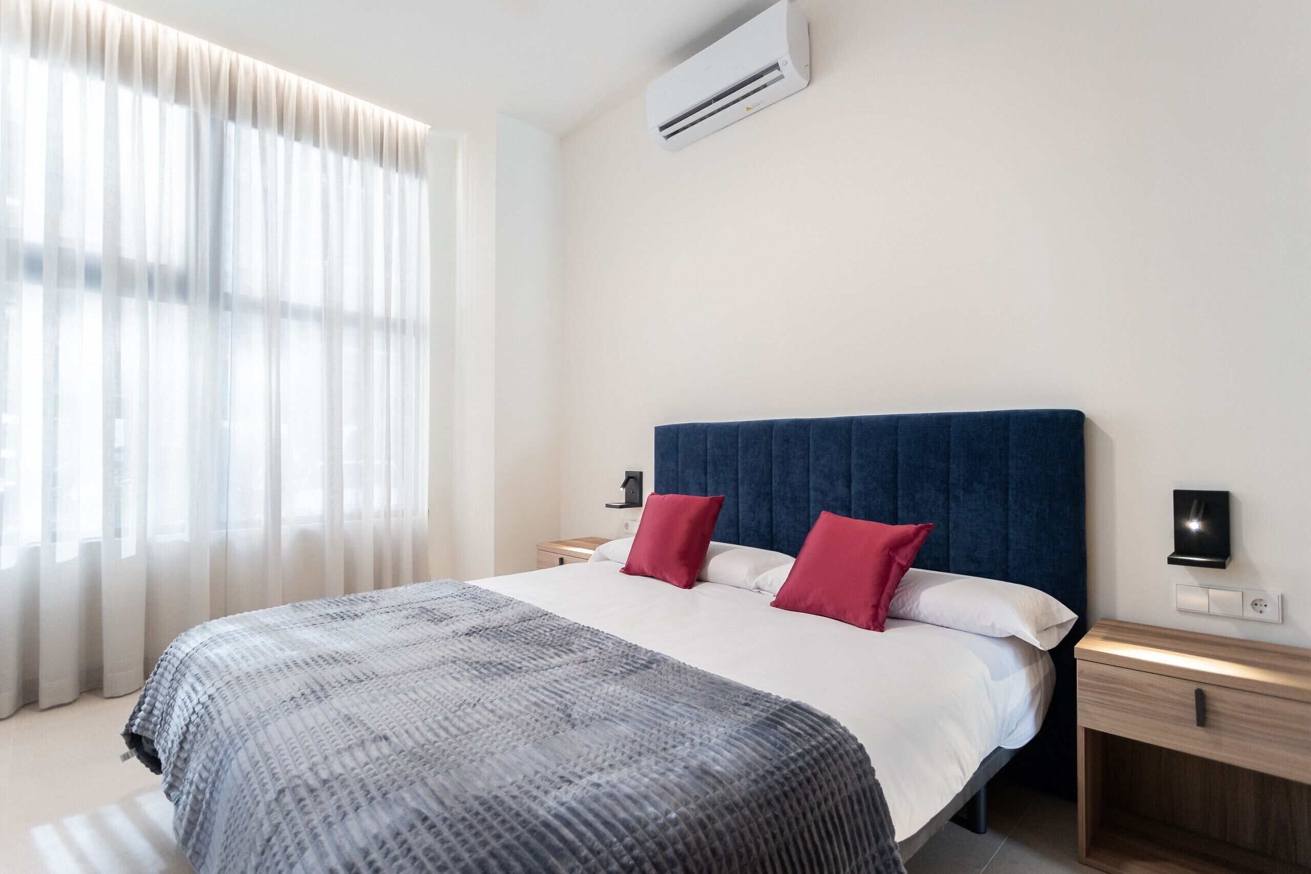 A modern hotel room with a large bed, a dark blue headboard, white bedding, and red pillows. There is a bedside table with a lamp and a wall-mounted air conditioner.