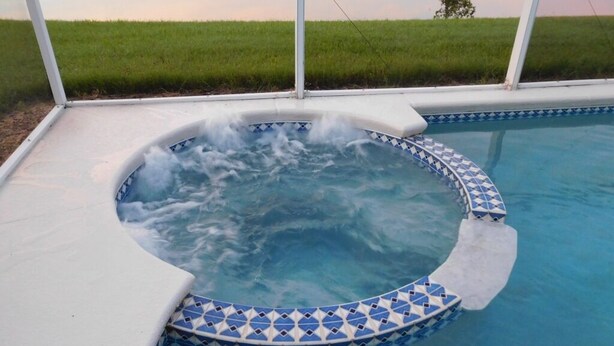 Un jacuzzi con borde de azulejos azules y blancos, rodeado por una terraza blanca, con un campo de césped al fondo.