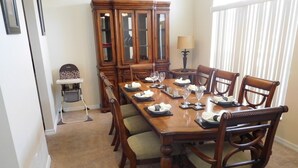 Un comedor con una larga mesa de madera dispuesta para seis comensales, rodeada de sillas a juego. Se observa una trona a la izquierda.