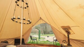 A tent with a bed, a hanging light fixture, a table with a kettle, and a view of the outdoors.