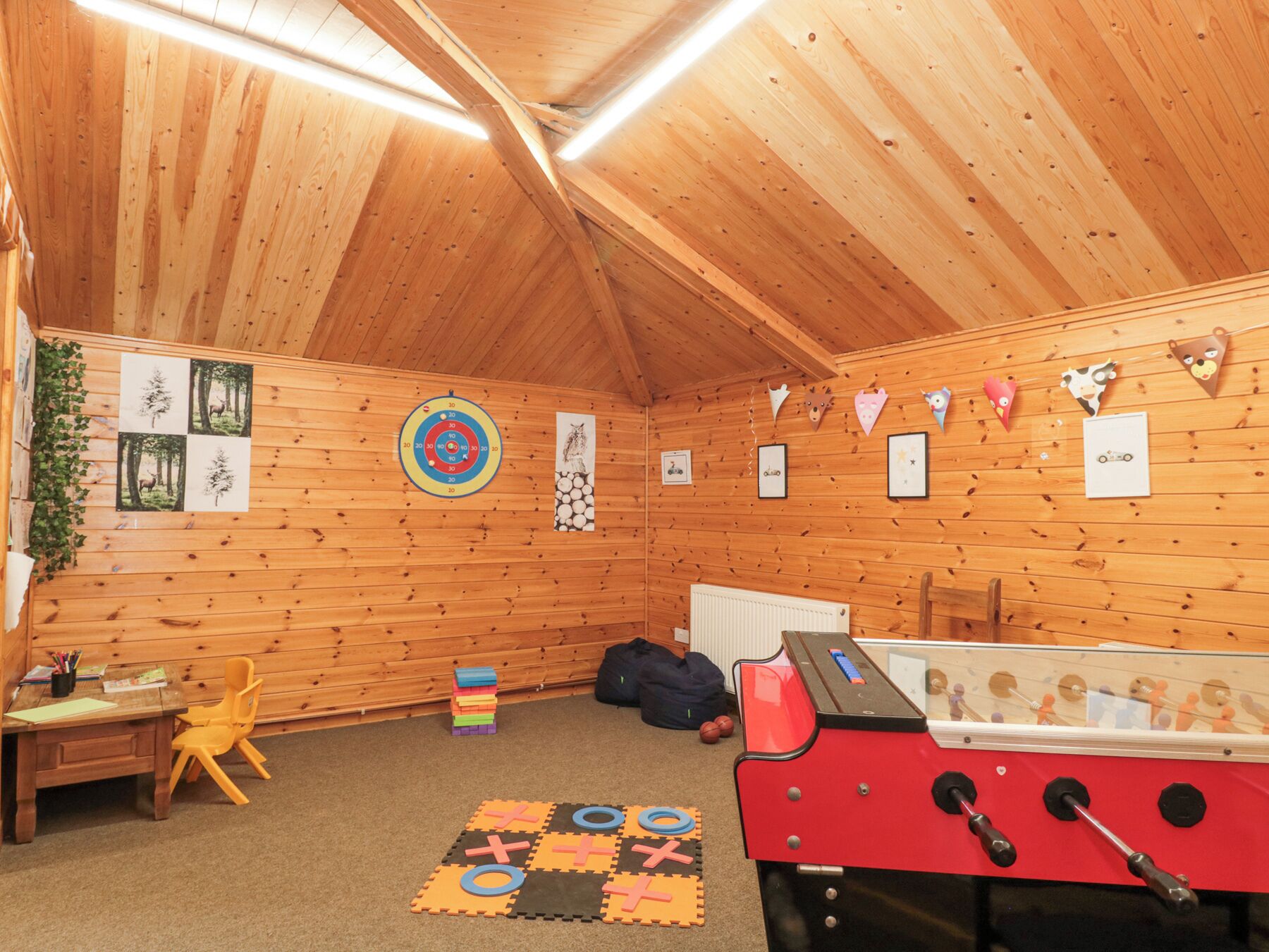 A cozy room with wooden walls, a foosball table, a dartboard, and a small desk with a chair.