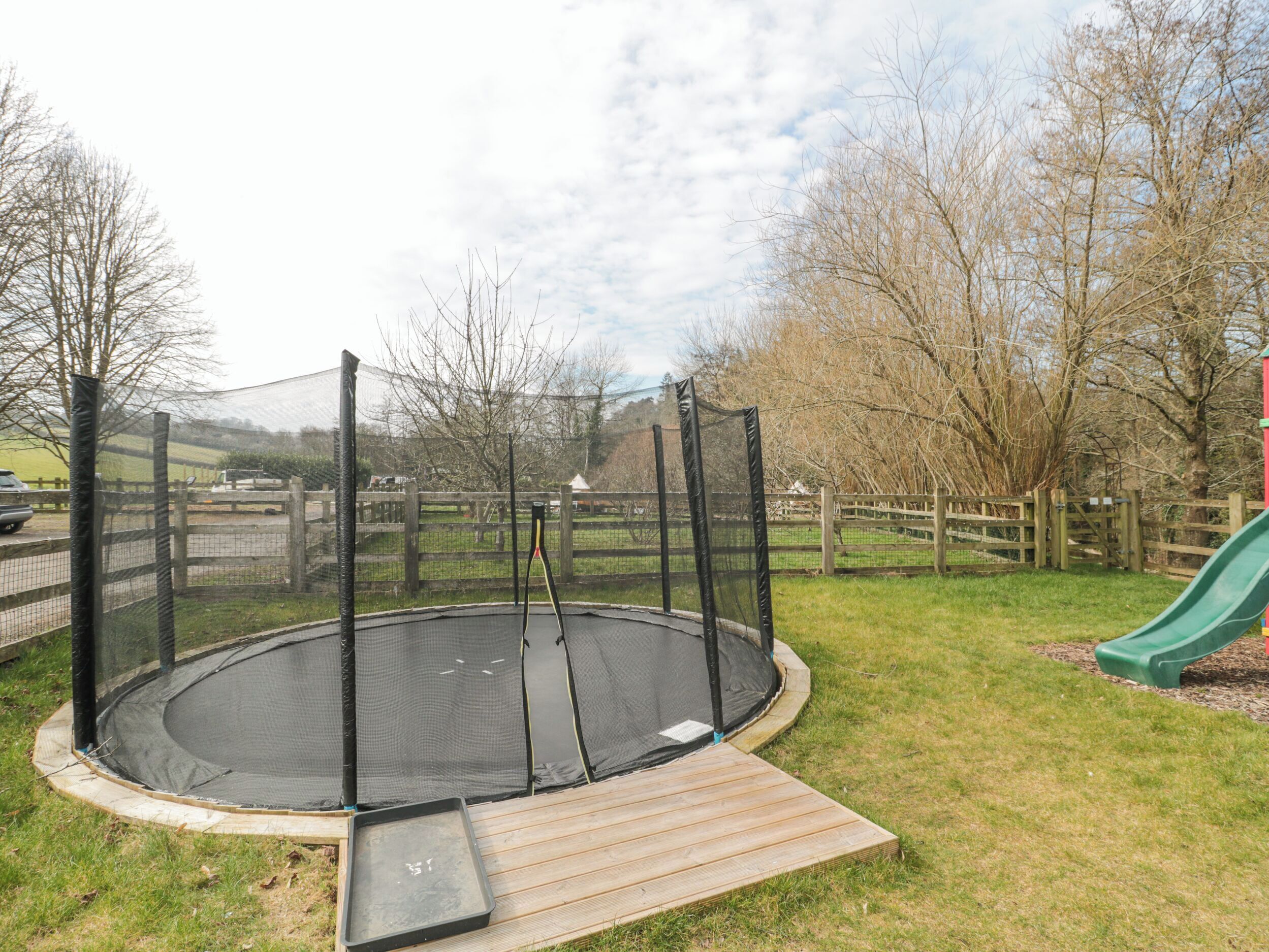 Ein Trampolin mit Sicherheitsnetz, umgeben von einem Holenzaun, in einem Rasenstück mit einer grünen Rutsche.