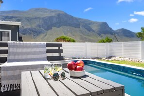 A poolside area with a wooden table, a towel, a bowl of fruit, and glasses of a beverage. - Metalasia Four - Three Bedroom House, Sleeps 6 (Betty's Bay)
