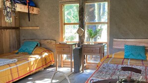A rustic cabin interior with two wooden beds, a wooden table, a kettle, and a window with a view of trees.
