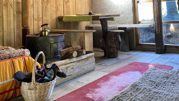 A rustic cabin interior with a wooden interior, a fireplace, a table with a kettle, and a window with a view of the outdoors.