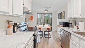 A modern kitchen with white cabinets, a central island, a dining area with a table and chairs, and a view through a glass door to the outside. - Remodeled! Beaverton 2BR/2BA Fenced Backyard Extended Stays (Beaverton)